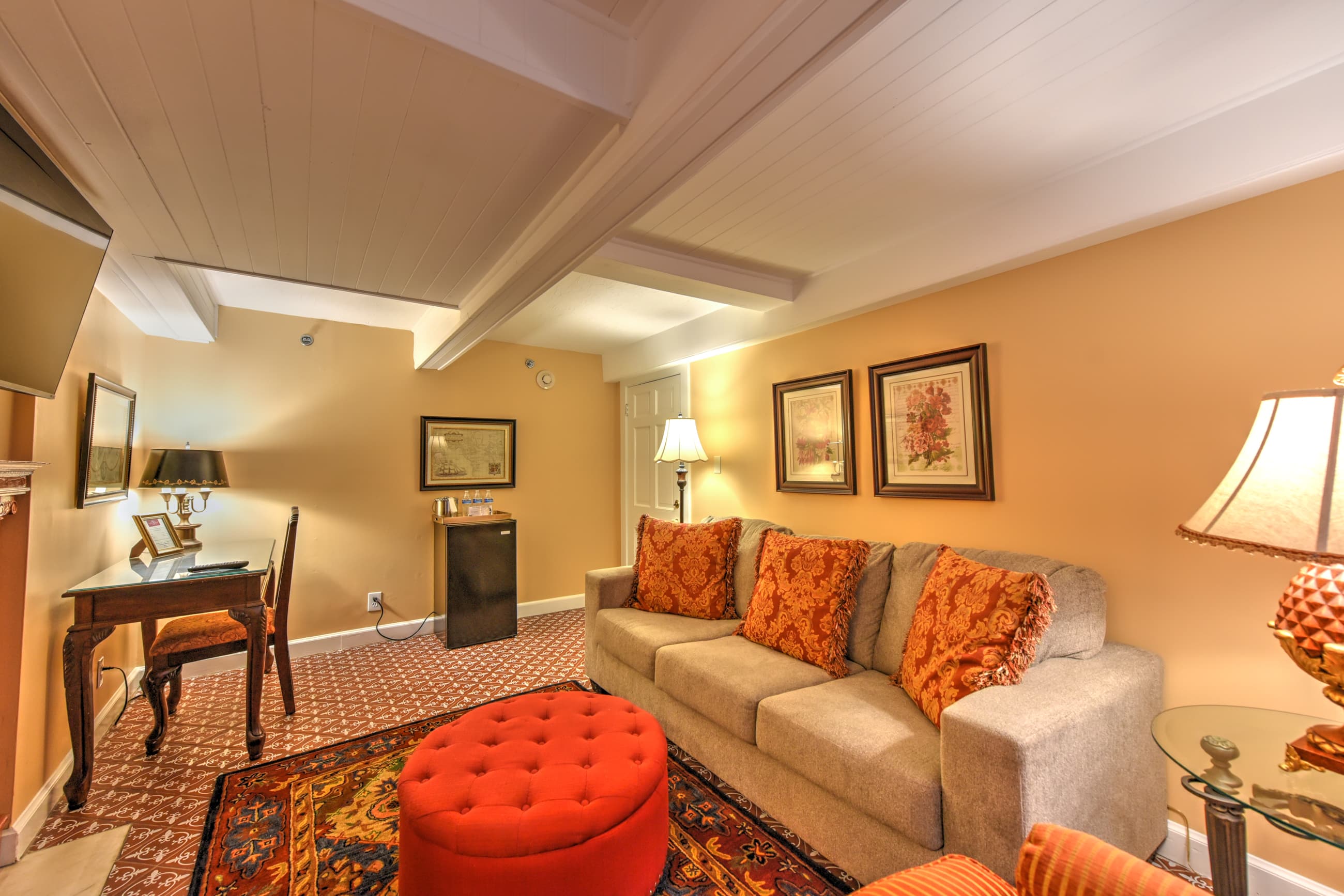 Cozy sitting area featuring a gray sofa with orange cushions, an ottoman, a lamp, and a small desk against warm-toned walls.
