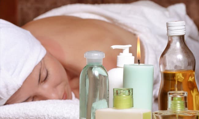 A woman relaxes with her back to the camera, surrounded by various skincare products and a lit candle.