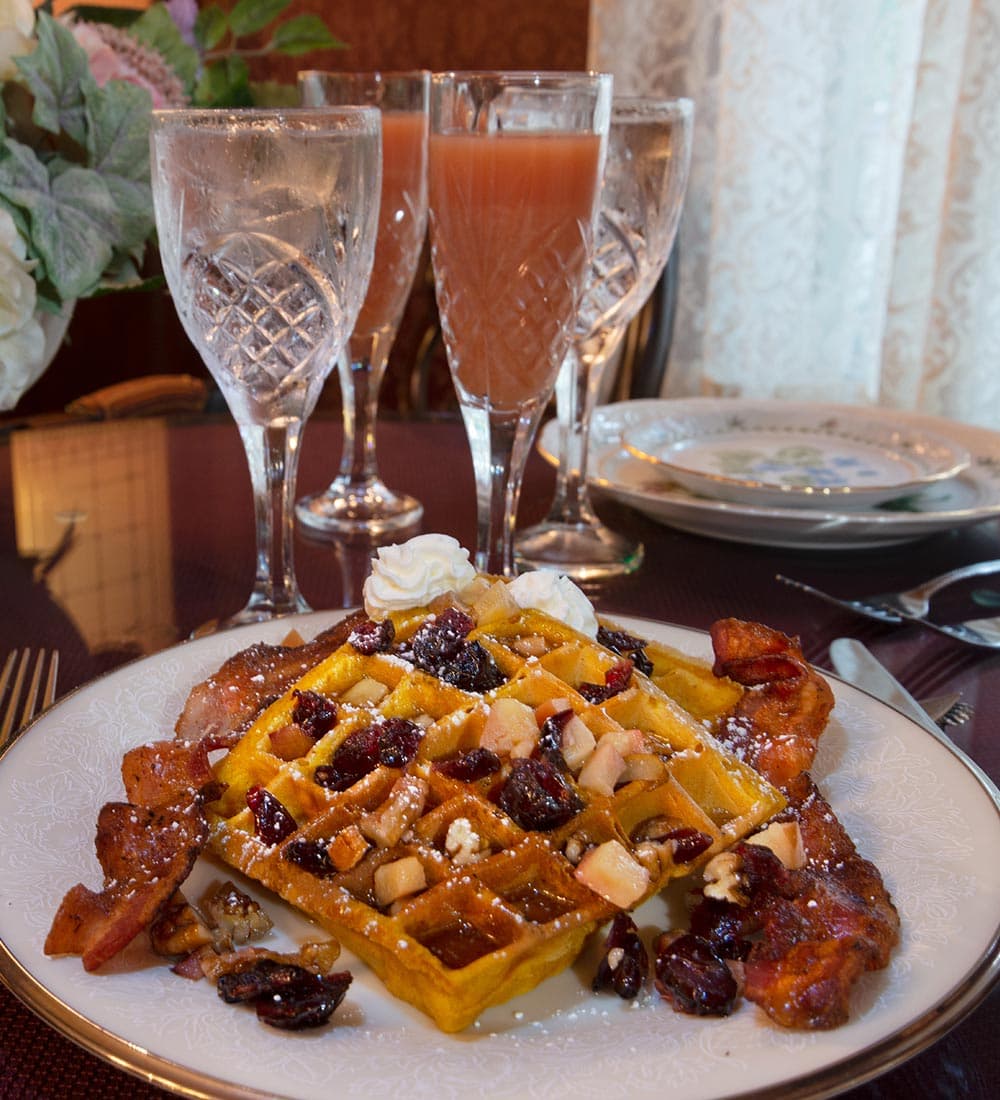 A plate of waffles topped with fruit, nuts, and whipped cream, served alongside bacon, with drinks in the background.