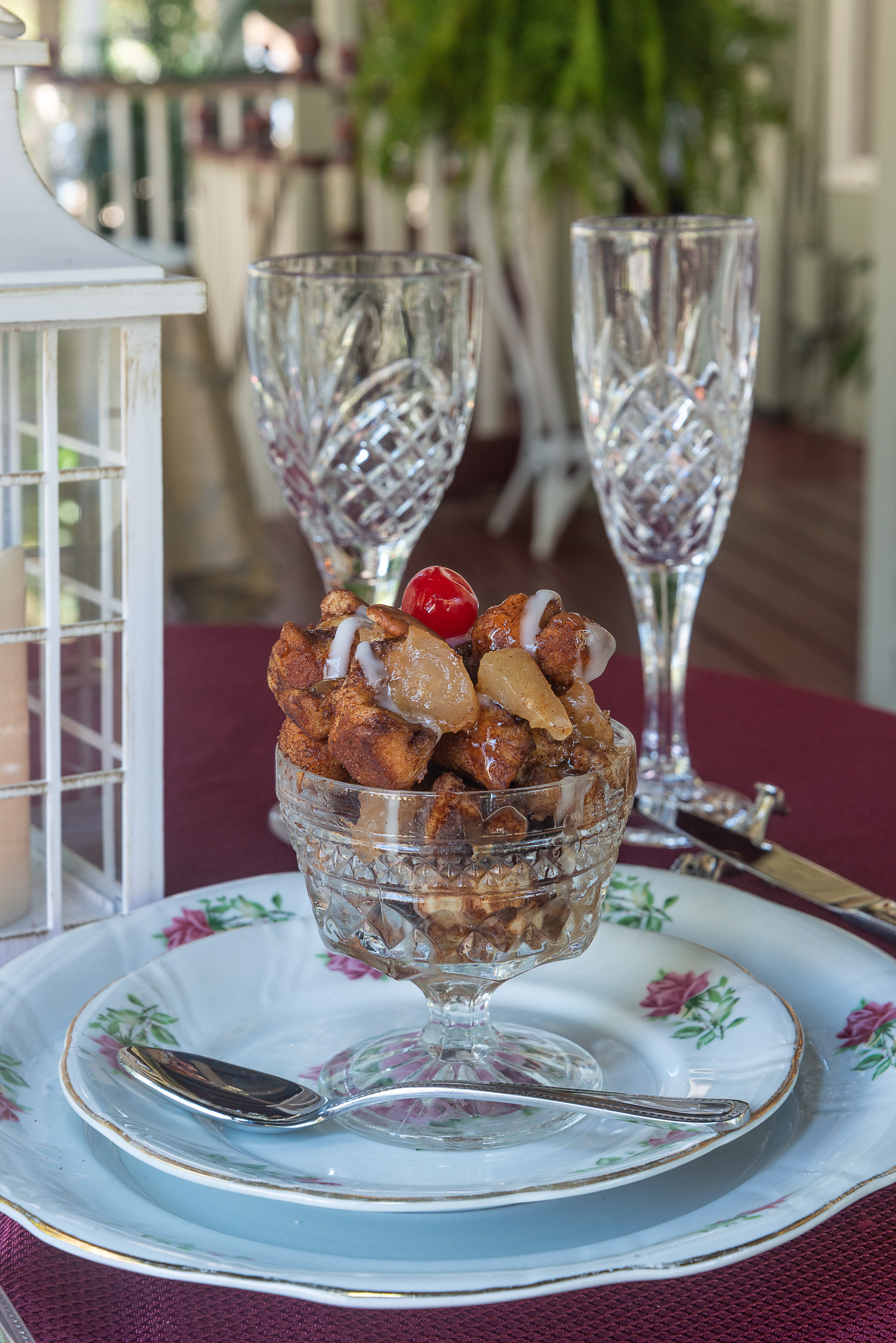 A dessert glass filled with cinnamon apple bread pudding topped with a cherry and drizzled with icing, set on a decorative table.