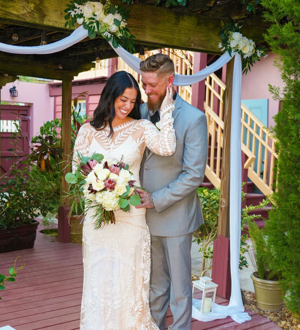 A joyful couple stands together under a floral arch, holding a bouquet, in a vibrant outdoor wedding setting.