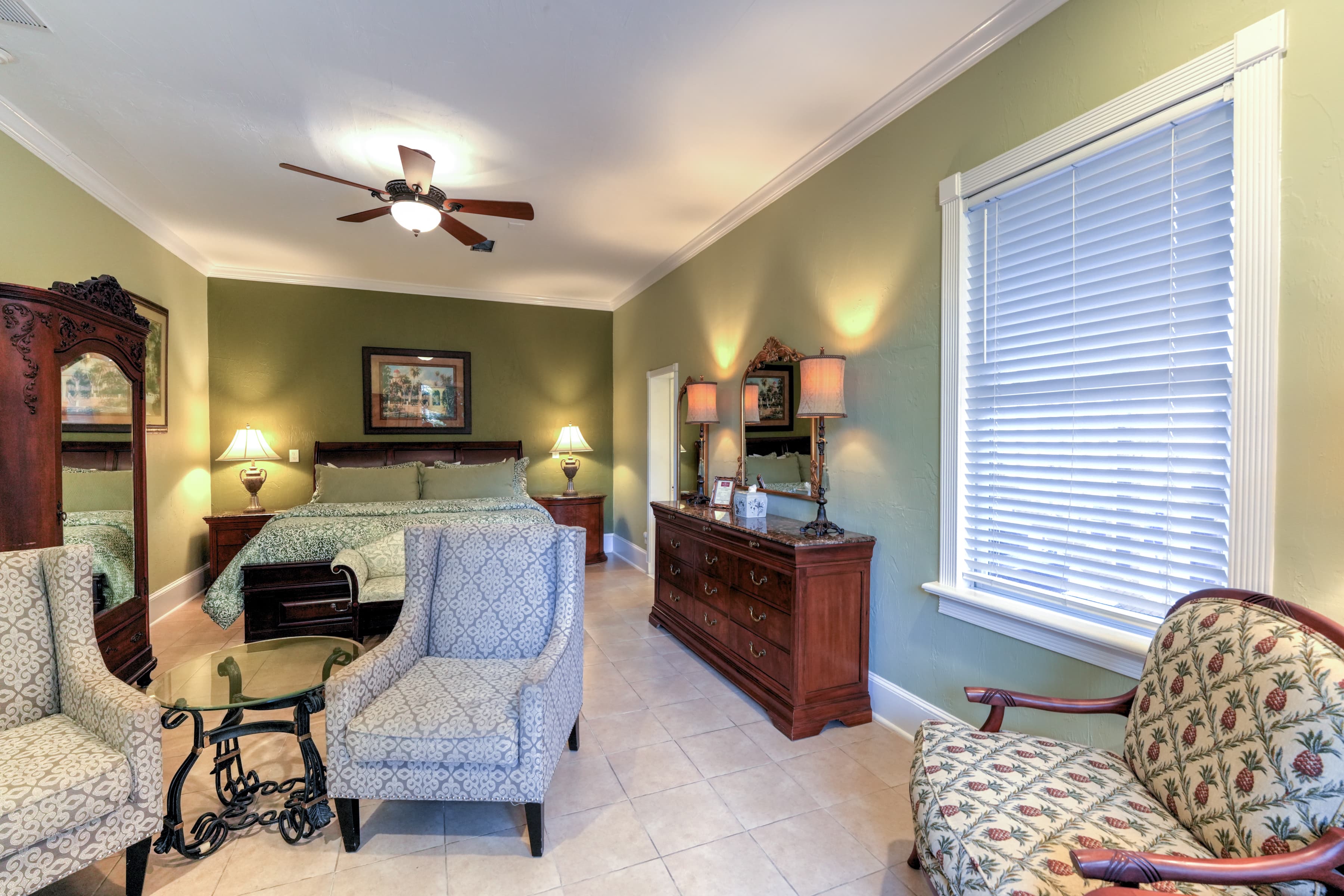 A cozy bedroom with a king-size bed, two upholstered chairs, a wooden dresser, and soft lighting against a green wall.