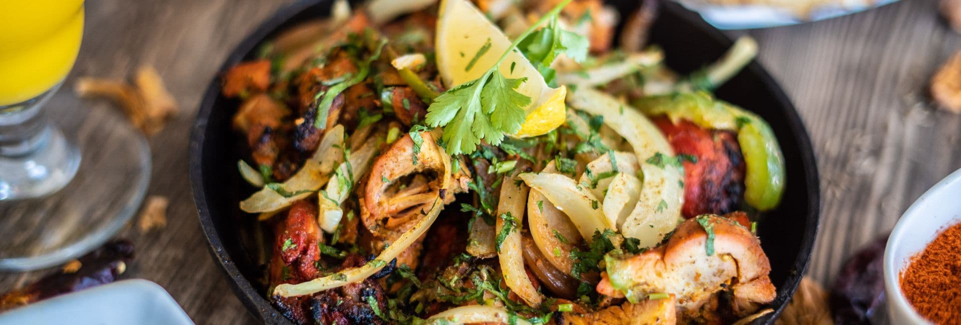 A sizzling skillet of grilled meat topped with onions, cilantro, and lemon wedges.
