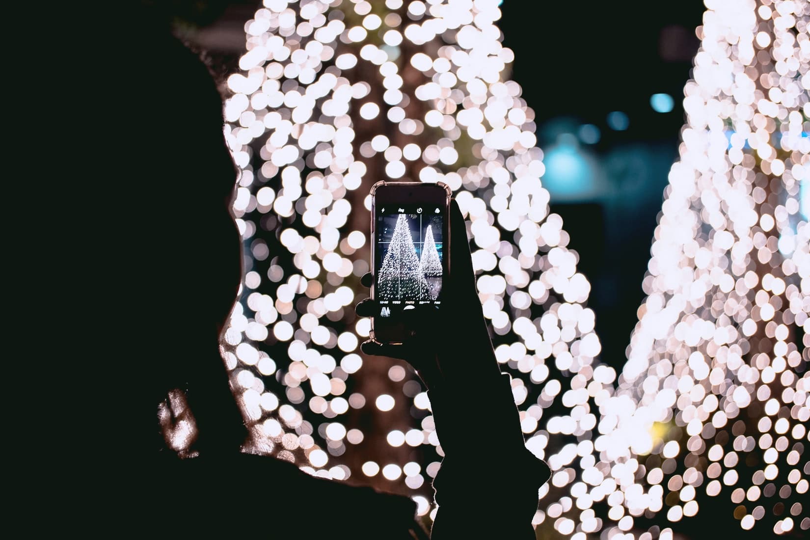 A person photographs a festive scene of illuminated trees with a smartphone, surrounded by soft bokeh lights.