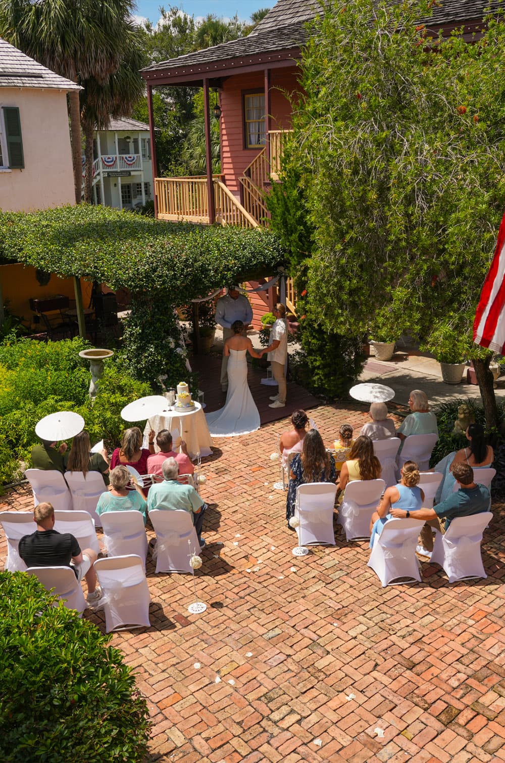 A bride and groom exchange vows in a garden setting, surrounded by guests seated on white chairs.