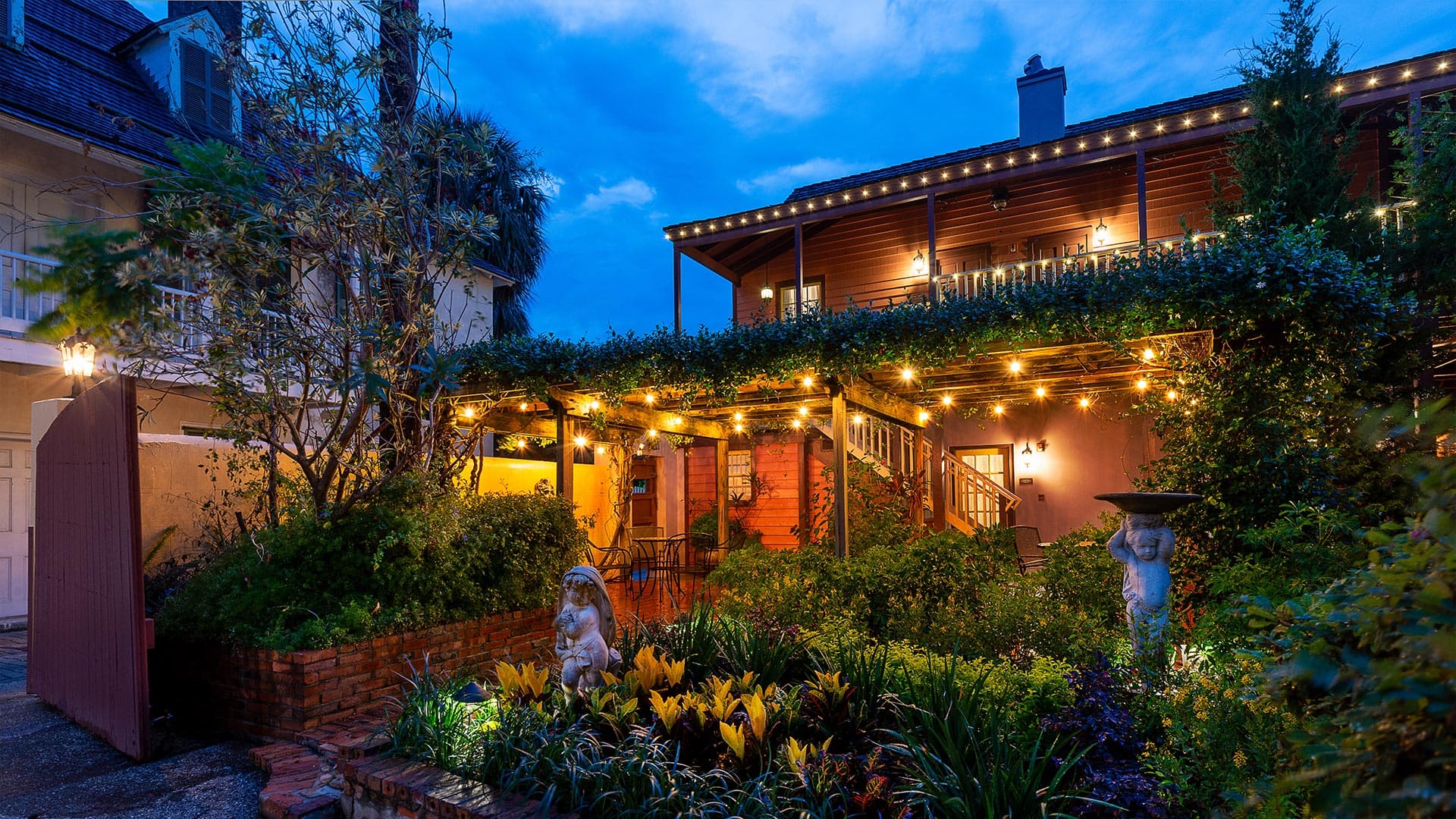 A tranquil outdoor space featuring lush greenery, a statue, and warm string lights against a twilight sky.
