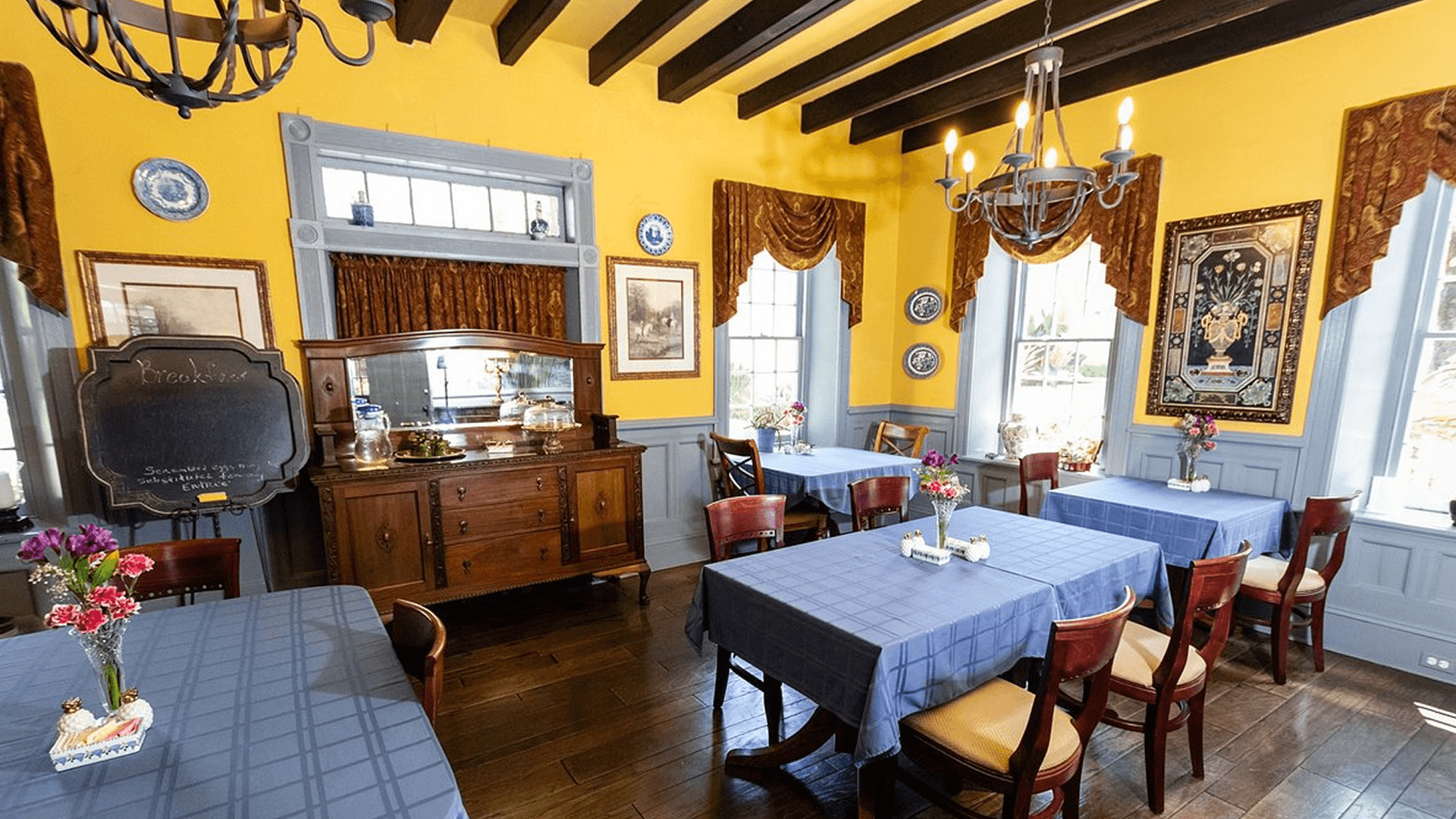 A cozy dining room with yellow walls, a wooden ceiling, and several tables set with blue tablecloths. There is a large wooden sideboard and a chalkboard in the background.