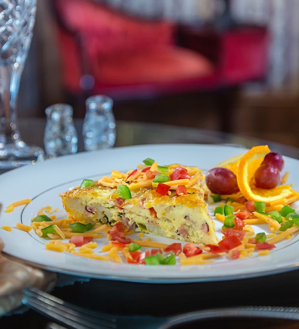 A slice of frittata topped with diced tomatoes, green peppers, and cheese, served with orange and grapes on a plate.