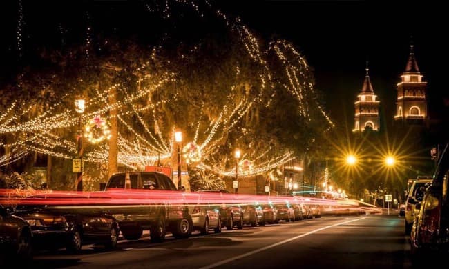 A street illuminated with festive lights and traffic at night, featuring two tall towers in the background.
