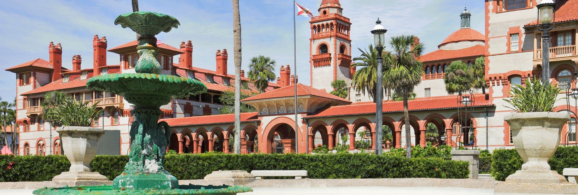 A decorative fountain in the foreground with a historic building featuring red roofs and palm trees in the background.