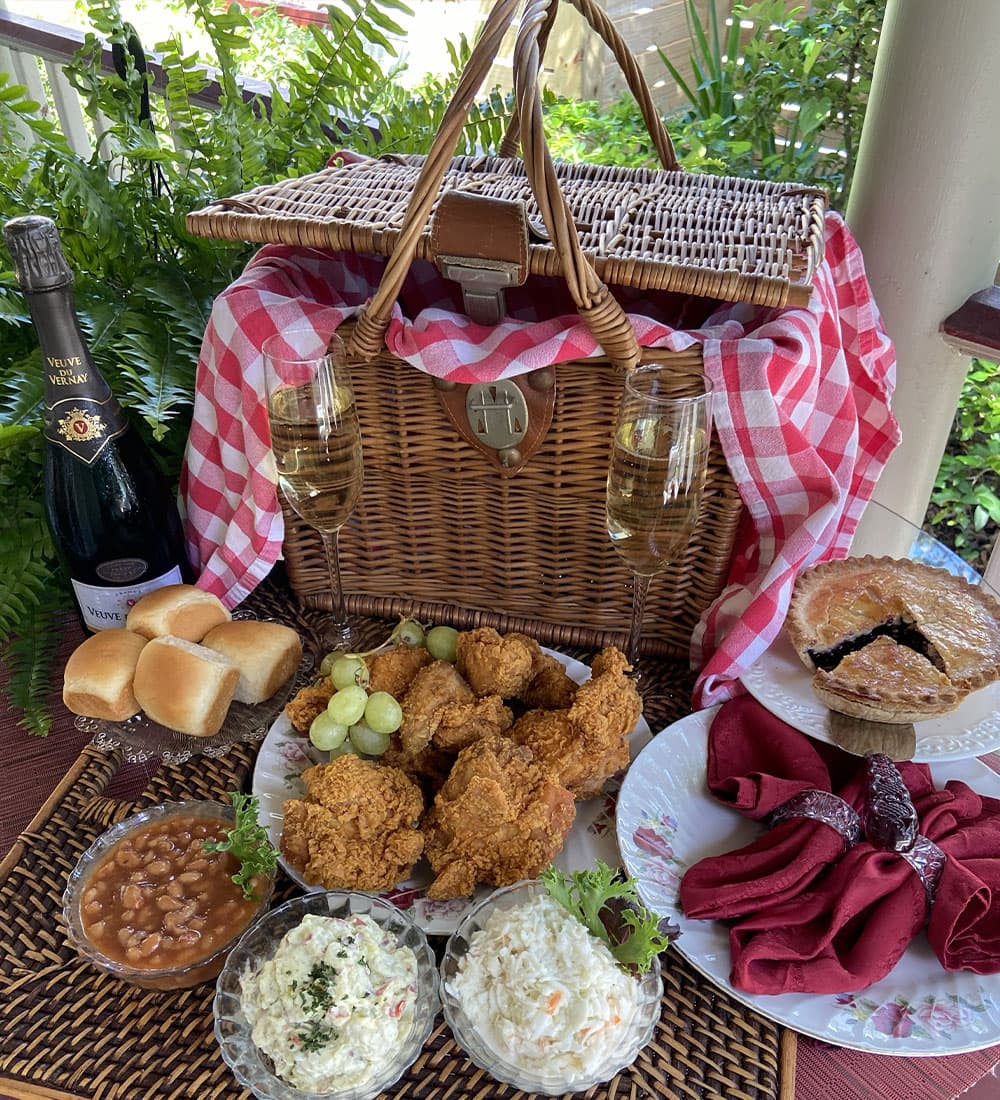 A picnic spread featuring fried chicken, sides, rolls, a bottle of champagne, and a pie, arranged around a wicker picnic basket.