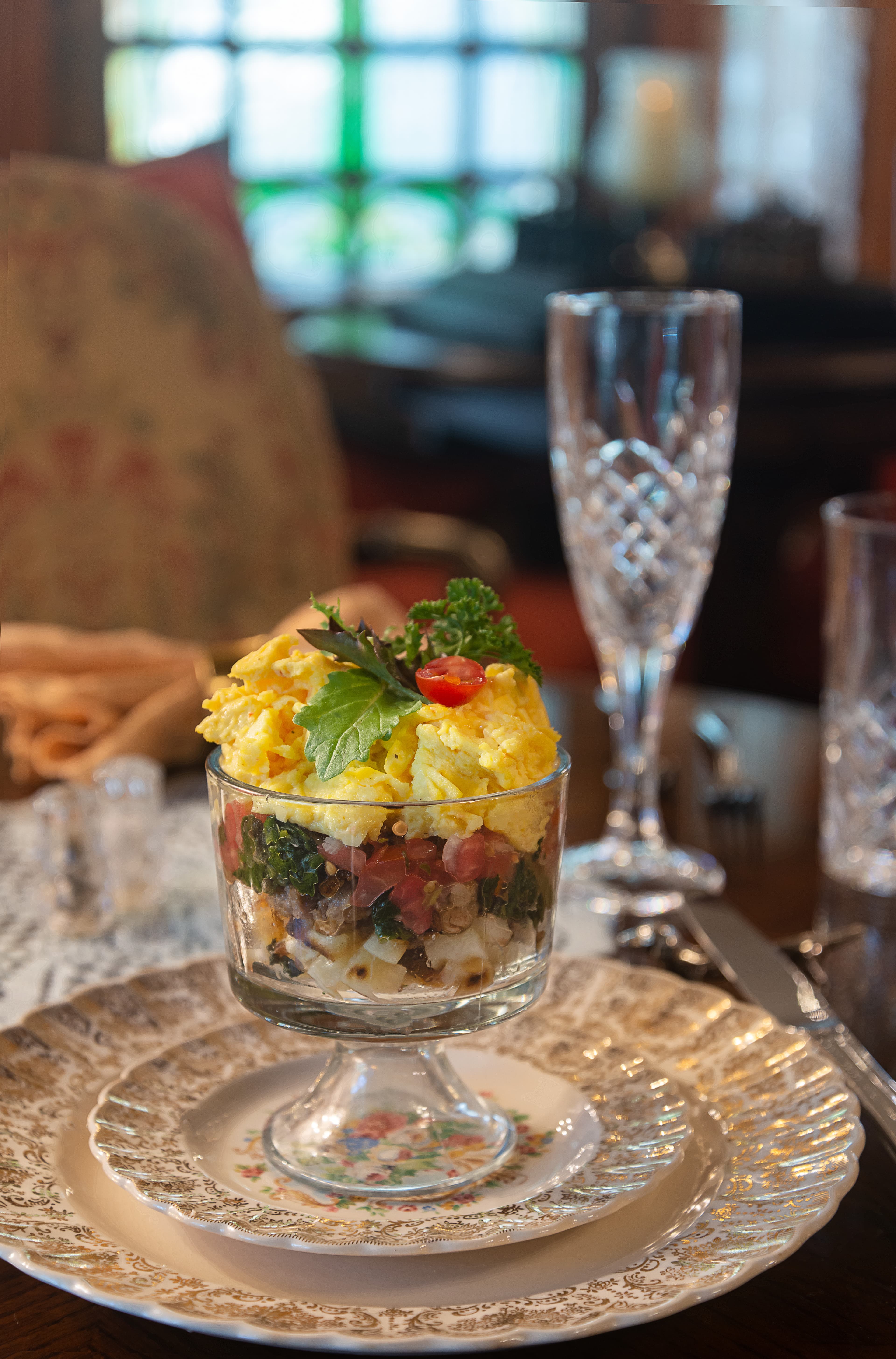 A layered dish in a glass featuring scrambled eggs, vegetables, and herbs, set on an ornate plate.