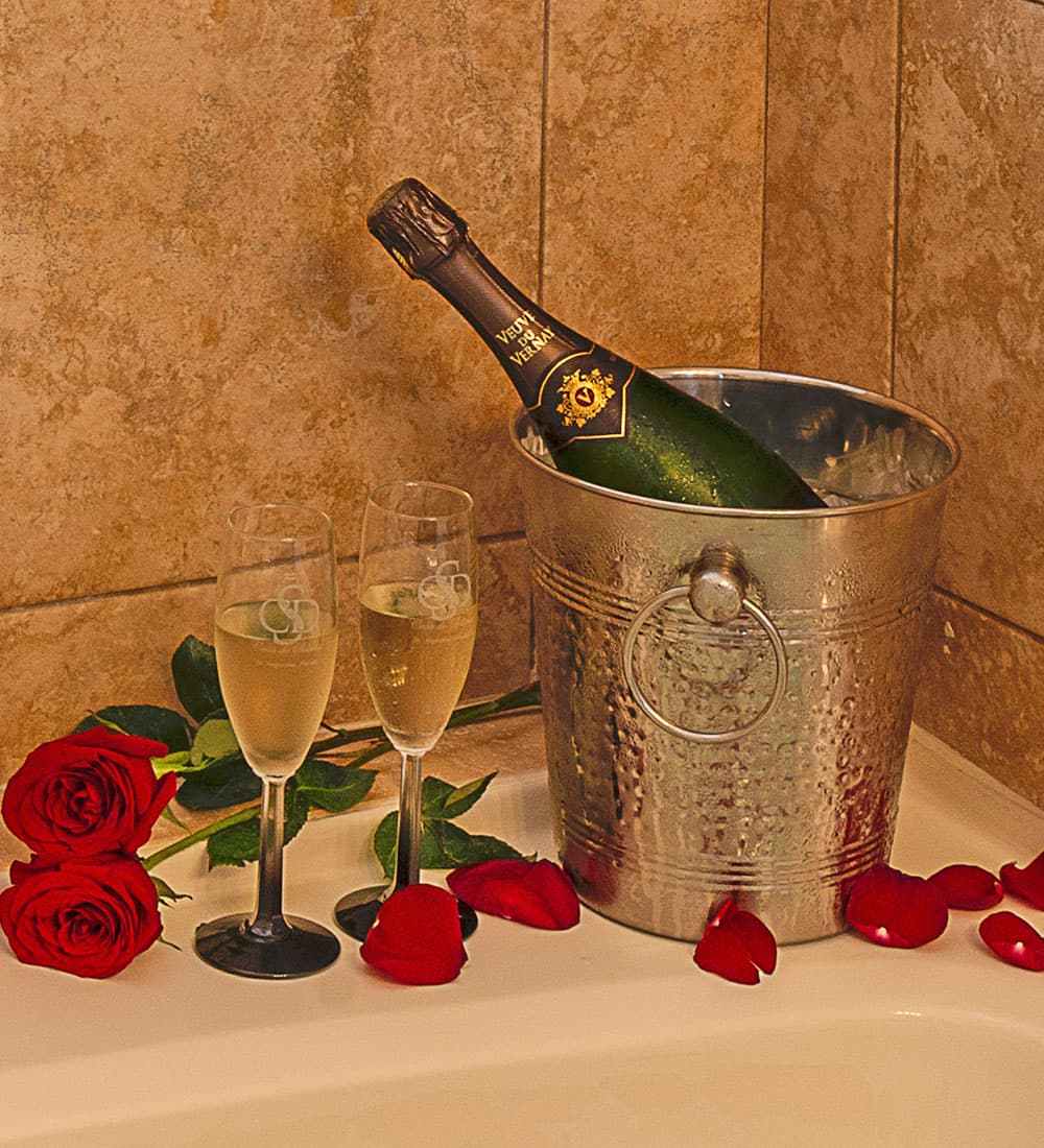 A bottle of champagne in an ice bucket, accompanied by two glasses and red roses.
