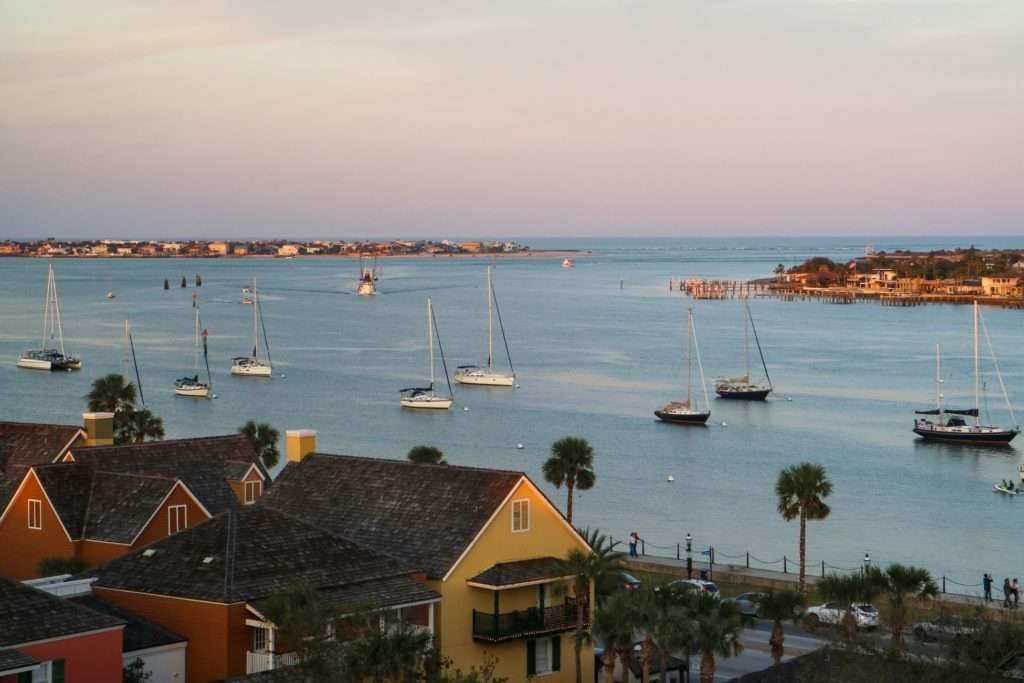 A view of sailboats on a calm bay at sunset, framed by colorful waterfront houses.