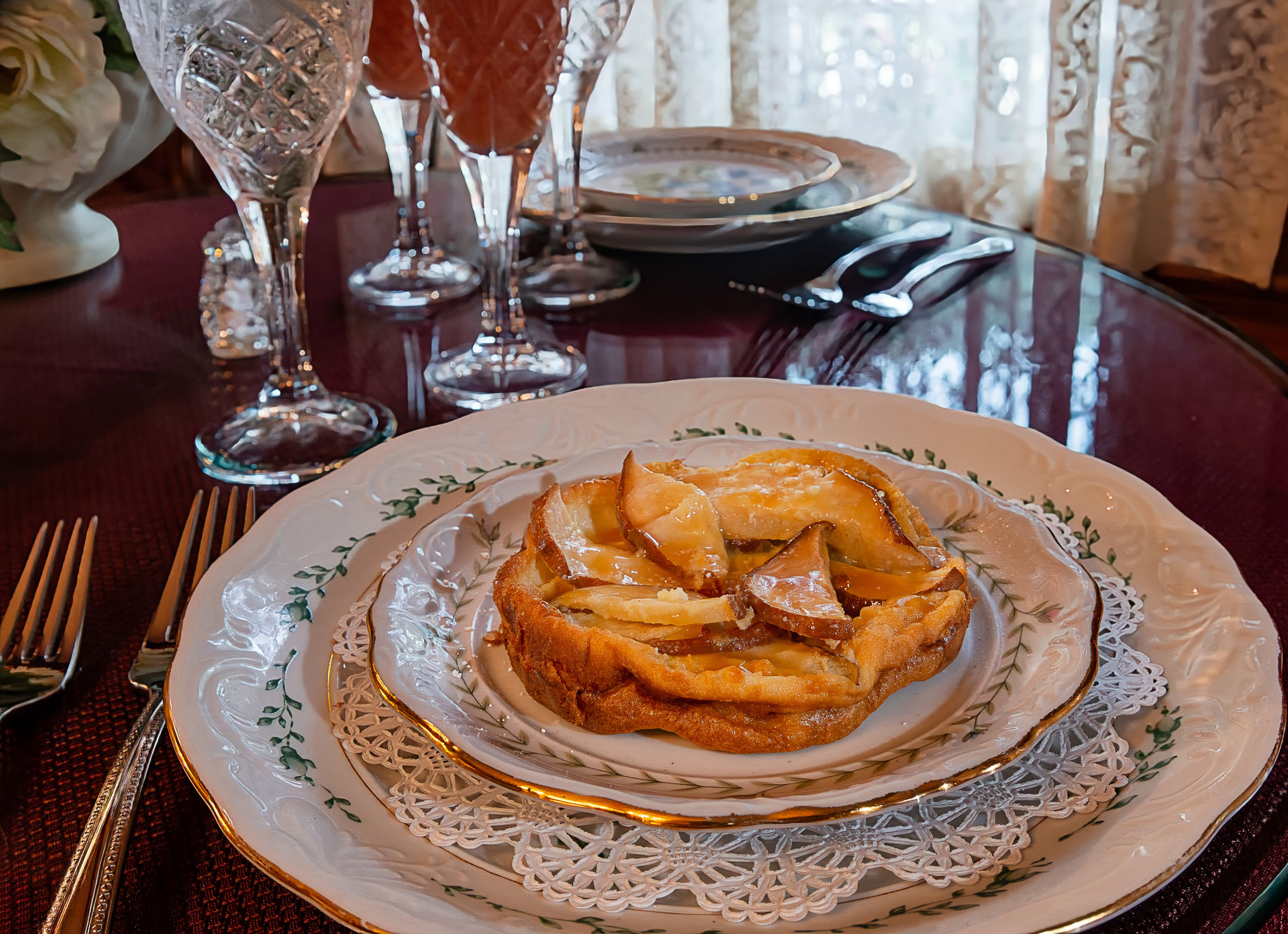 A plate of apple dessert sits elegantly on a white lace doily, surrounded by crystal glasses and silverware on a richly colored table.