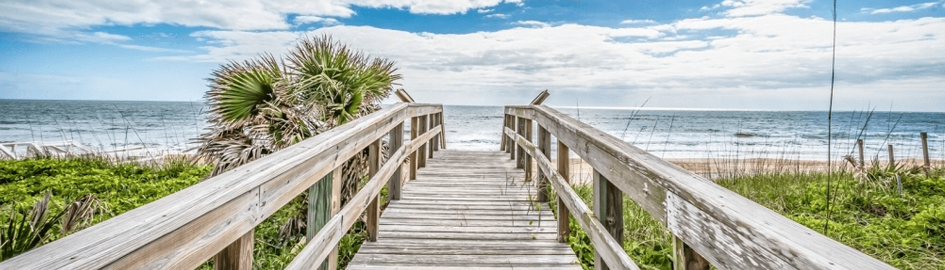 A wooden boardwalk leads to a sandy beach with a clear blue ocean and a bright sky.
