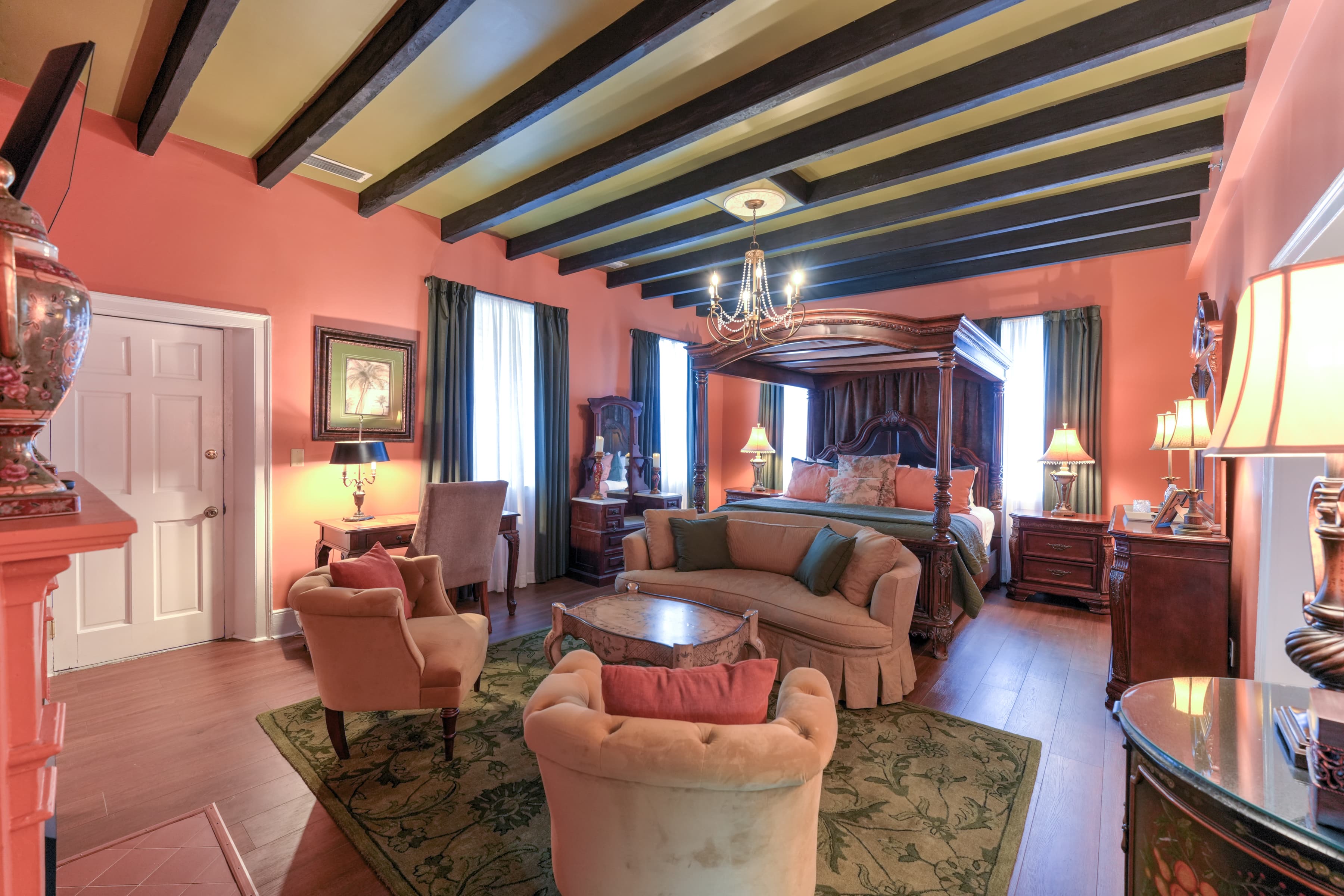 Romantic, luxurious guestroom with canopy four-poster king bed, beam ceiling, and sitting area with oriental area rug and upholstered loveseat and two chairs. A doorway leads to the private balcony with outdoor sitting area.