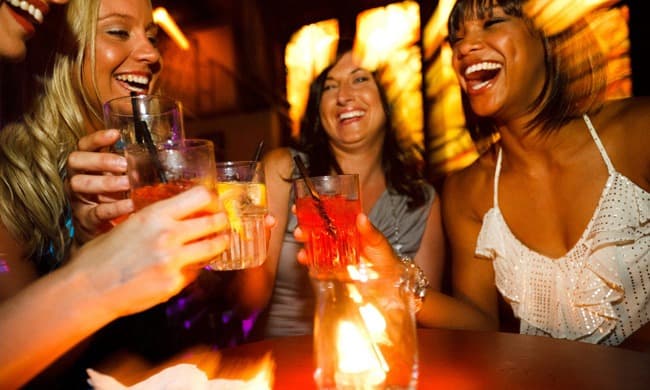 A group of four women joyfully clinks glasses while laughing in a dimly lit bar.