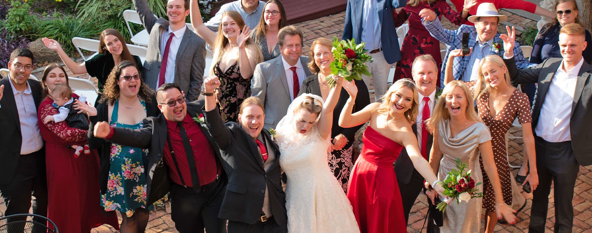 A joyful wedding party poses and celebrates outdoors, with the bride holding a bouquet at the center.