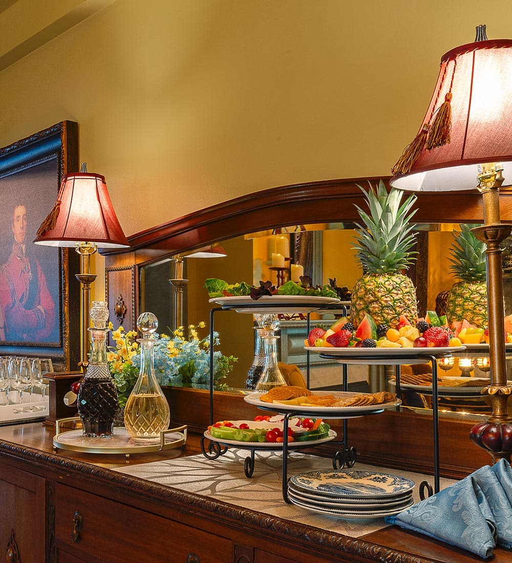 A decorative buffet table with fruit platters, drinks, and candles, set against an ornate mirror and a framed portrait.