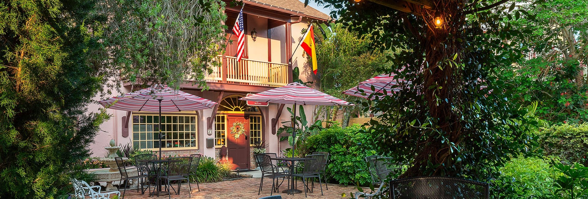 A charming courtyard featuring striped umbrellas and lush greenery in front of a building adorned with flags.