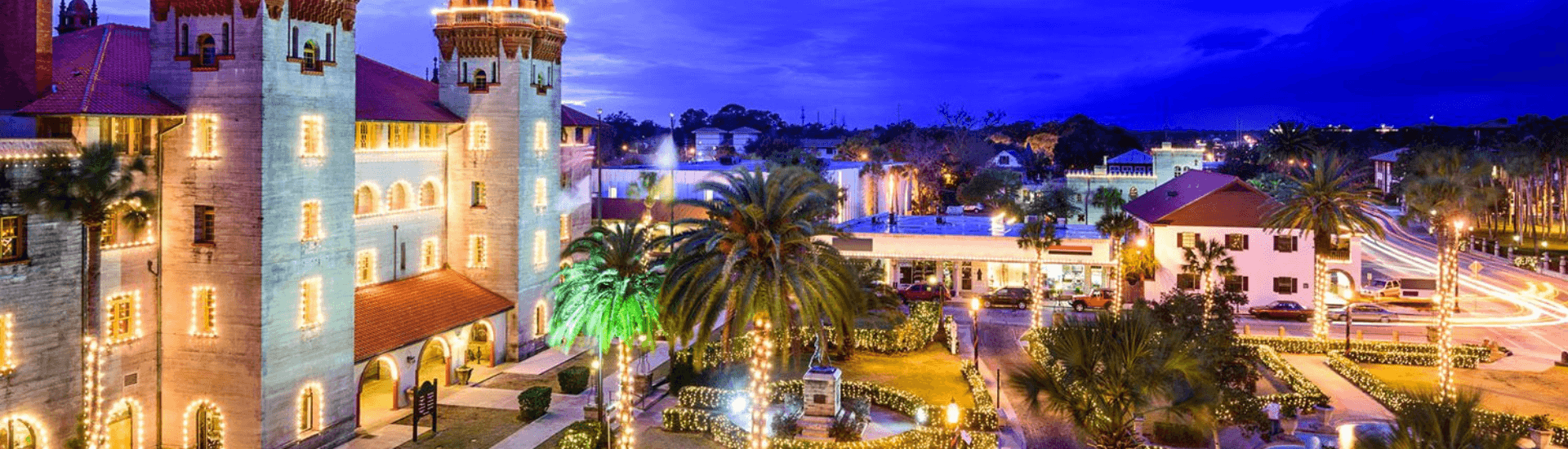 A night view of St. Augustine, Florida, during the Nights of Lights festival. The iconic Lightner Museum, adorned with Christmas lights, is the focal point of the image. Other buildings and streets are also illuminated, creating a festive atmosphere.
