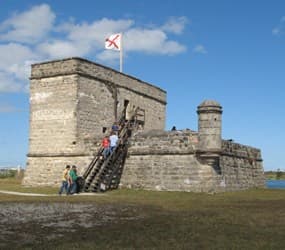 A historic stone fort with a flag, surrounded by grassy land and visitors ascending a wooden staircase.
