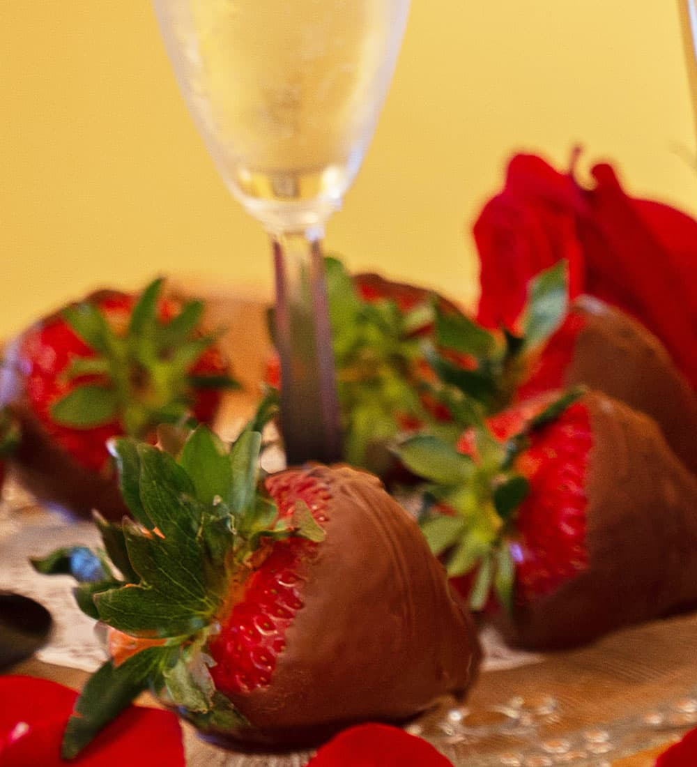 Chocolate-covered strawberries with a glass of sparkling wine and red rose petals arranged on a silver platter.
