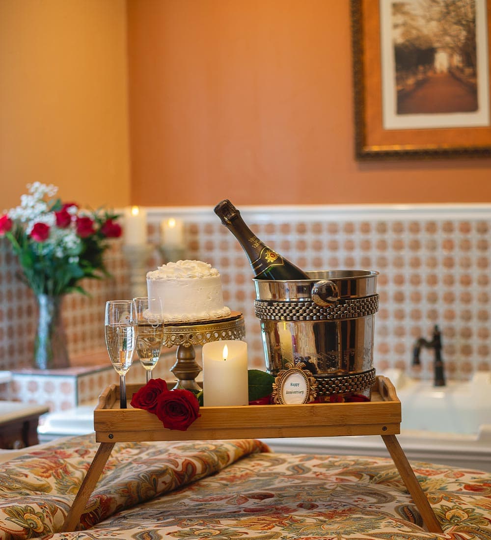 A romantic setup featuring a cake, champagne in a cooler, glasses, and roses on a wooden tray.
