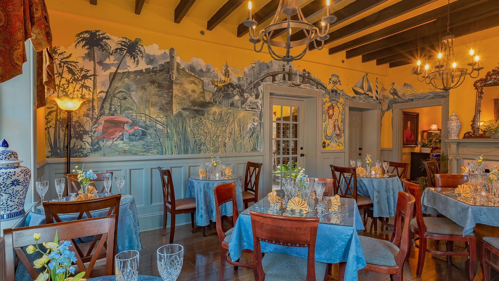 A cozy dining room featuring blue tablecloths, wooden chairs, and vibrant murals on the walls.