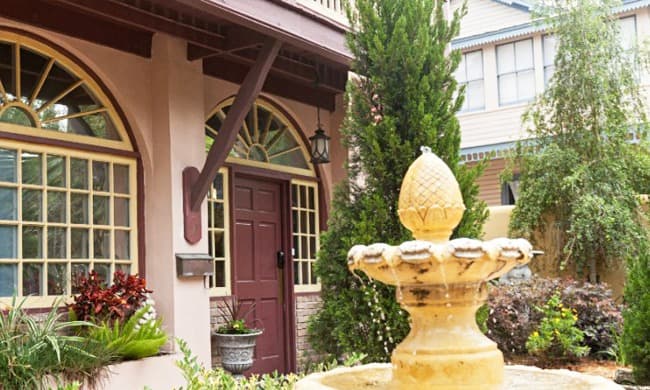 A fountain with a decorative top is situated in front of a charming house with large windows and a dark red door.