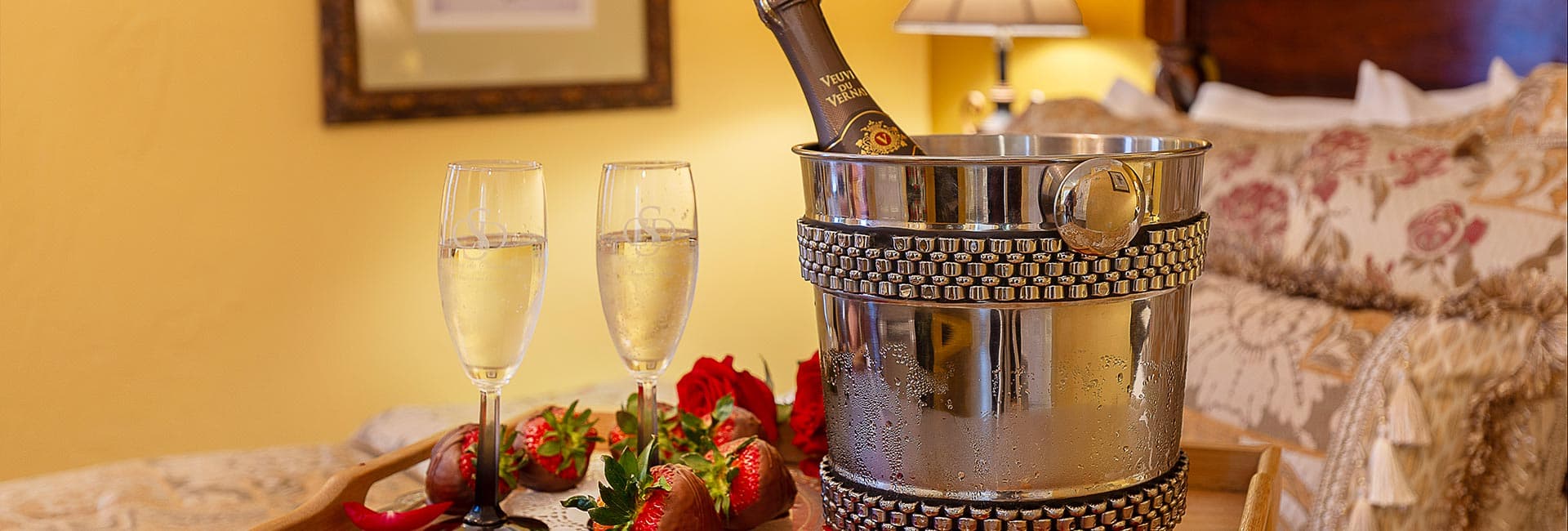 A silver ice bucket with champagne, two flutes, chocolate-covered strawberries, and red roses on a wooden tray.