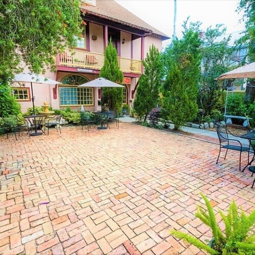 A brick patio surrounded by greenery and outdoor seating near a colorful building. A brick patio surrounded by greenery and outdoor seating near a colorful building.