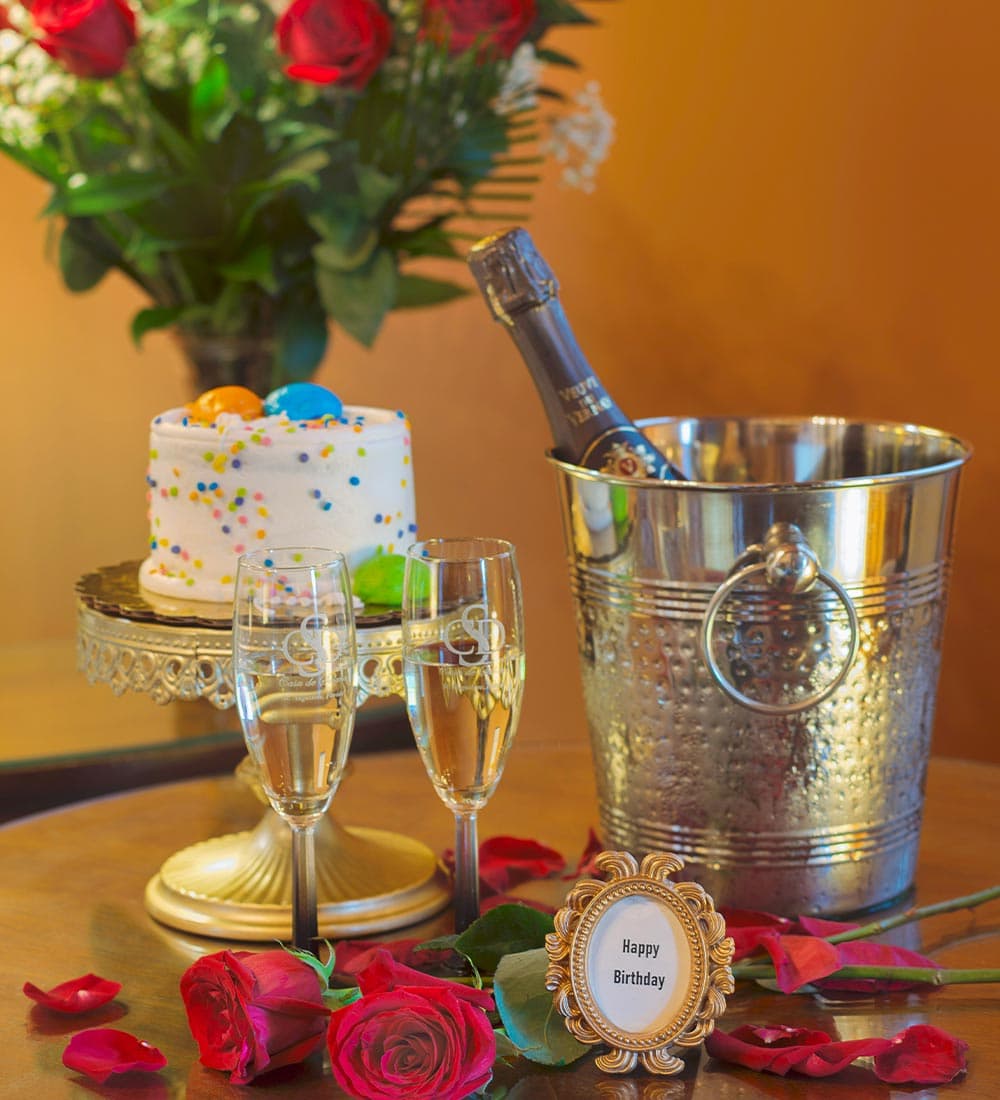 A birthday celebration setup featuring a cake, champagne, glasses, roses, and a "Happy Birthday" sign.