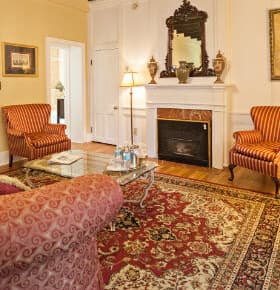 A cozy sitting room featuring two striped armchairs, a plush sofa, a glass coffee table, and a fireplace with a mirror above.