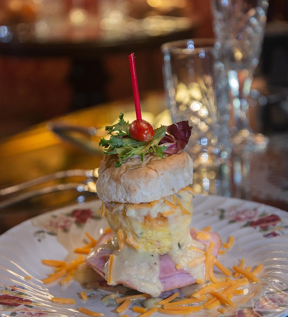 A layered dish featuring a biscuit topped with egg, ham, cheese, and garnished with greens and a cherry tomato.