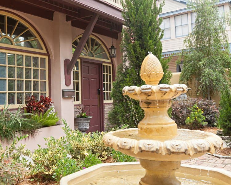 A decorative stone fountain with a pineapple finial is situated in a landscaped courtyard next to a charming building.