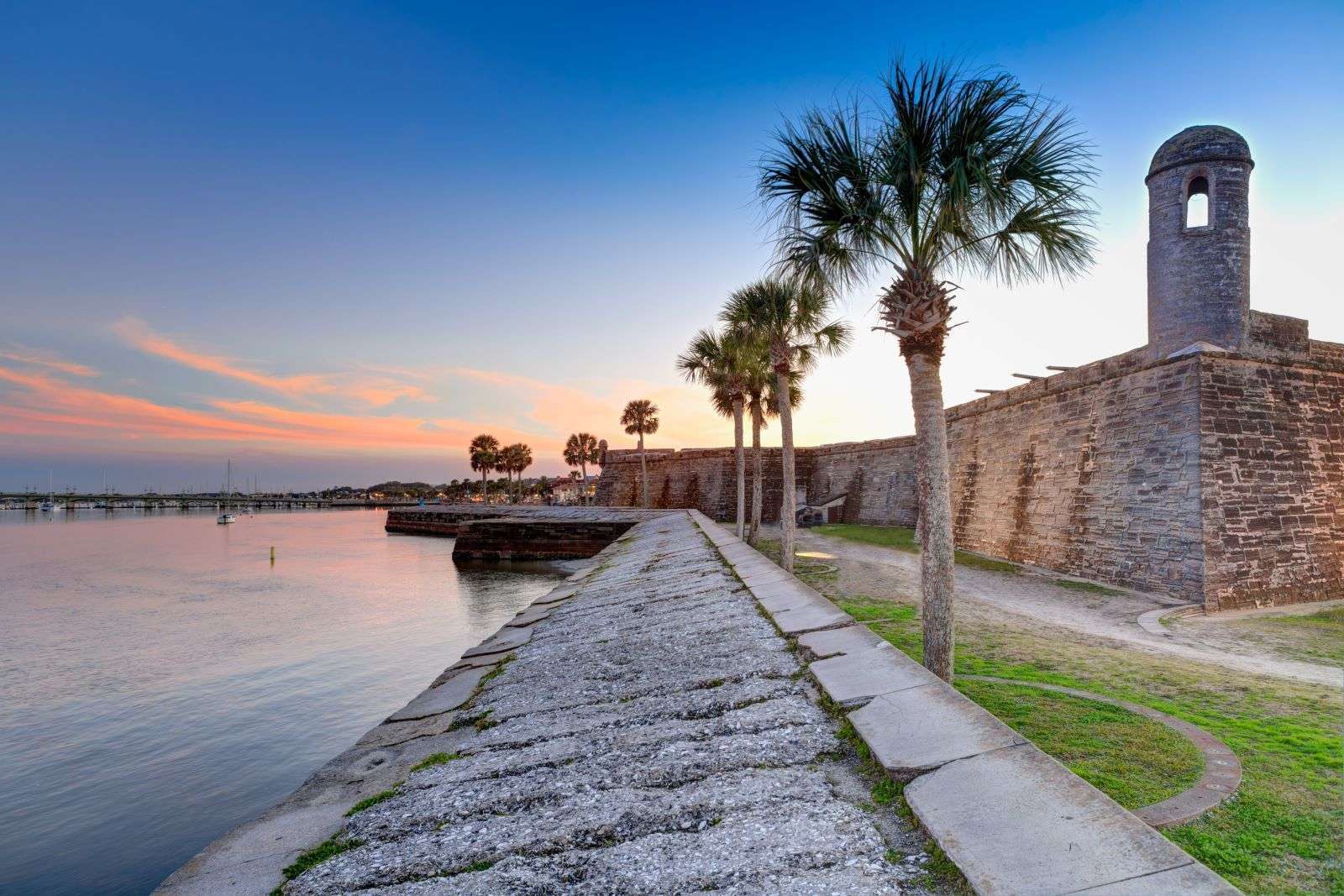 A stone pathway lined with palm trees leads to a historic fort beside a calm body of water at sunset.