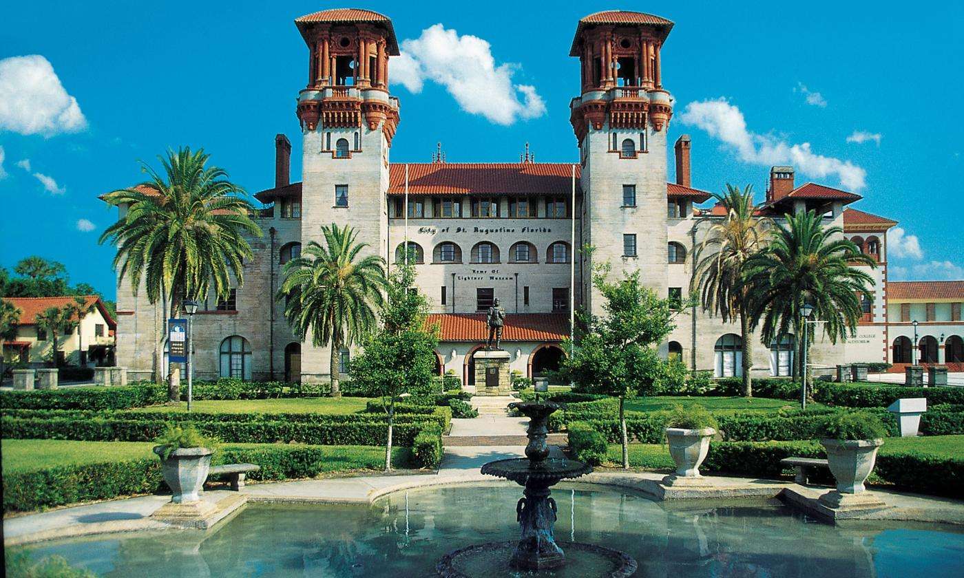 A grand historic building with twin towers surrounded by lush palm trees and a fountain in the foreground.