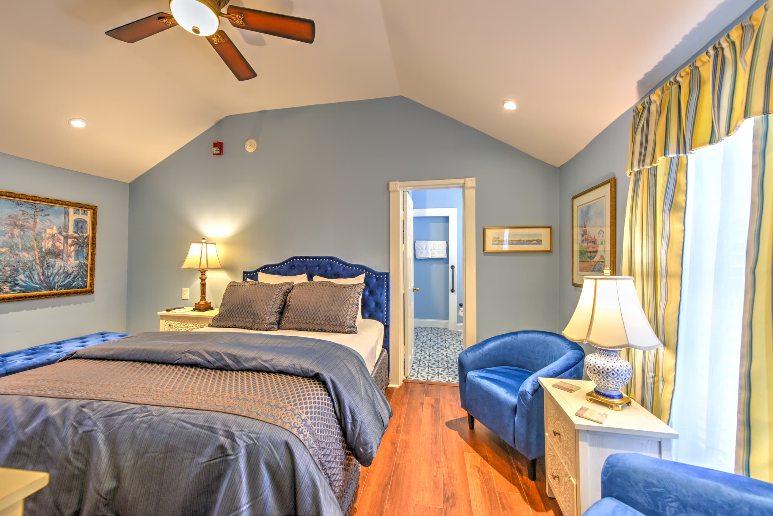 A queen bed with blue upholstered headboard with matching bedspread is the focal point of a bright, cheerful room. A large window allows lots of natural light into the room. Next to the bed is the doorway to the private bath