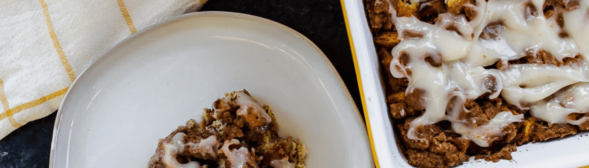 A plate of cinnamon roll French toast casserole with a drizzle of cream cheese frosting. The casserole is in a baking dish in the background.