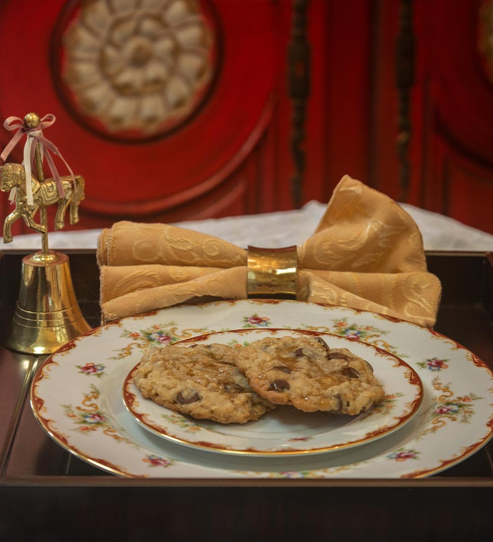 A decorative tray holding a plate of chocolate chip cookies, accompanied by a golden bell and a folded napkin.