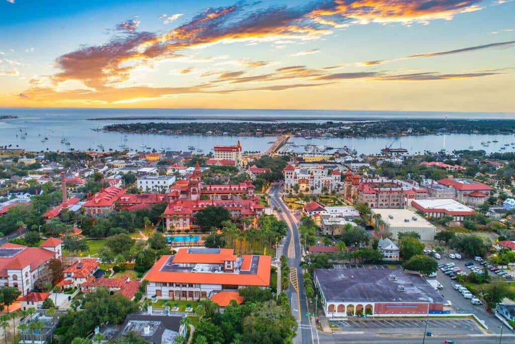 Aerial view of a coastal town at sunset, showcasing buildings with red roofs and a harbor in the background.