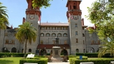 Green hedges and statue on pedestal in front of grey stone building with arched windows, two towers with red roofs